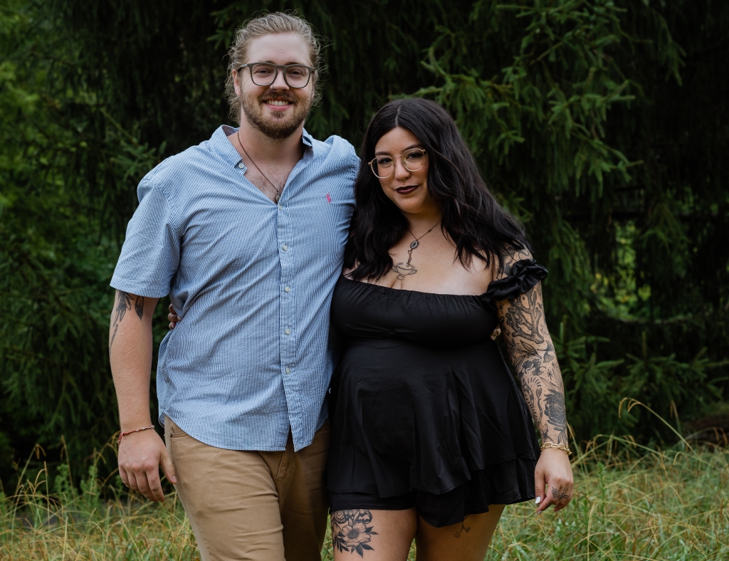 couple with tattoos posing arm in arm in front of a pine tree